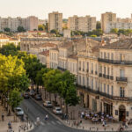 A high-angle view of a sunlit Nîmes street with historic buildings, cafes, trees, and people, contrasting with distant modern apartments.